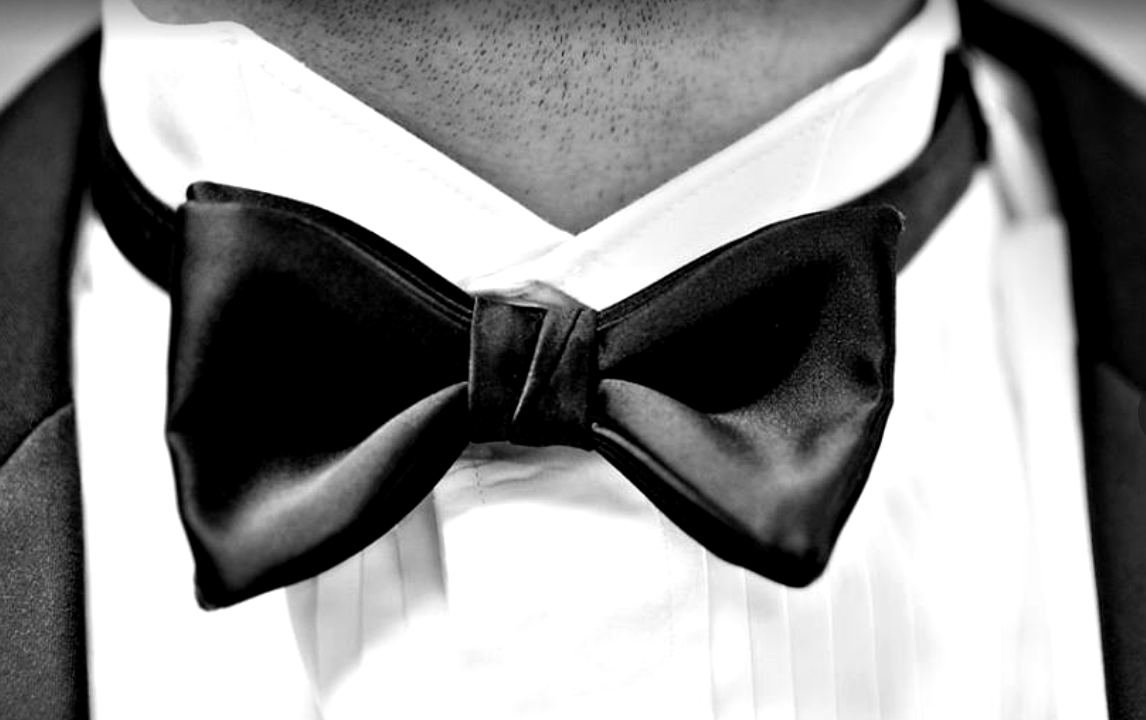 A black satin bow tie, tied, at the collar of a white pleated tuxedo shirt, in black and white.
