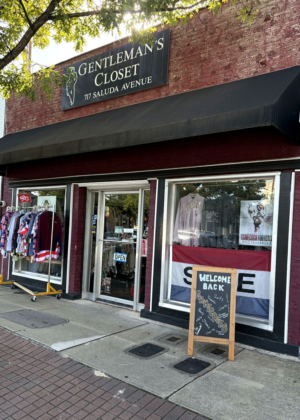The Gentlemen's Closet storefront on Saluda Avenue — dark red brick, a black awning, the serif sign with its gentleman silhouette, a sidewalk rack of shirts, and a chalkboard listing what is inside.