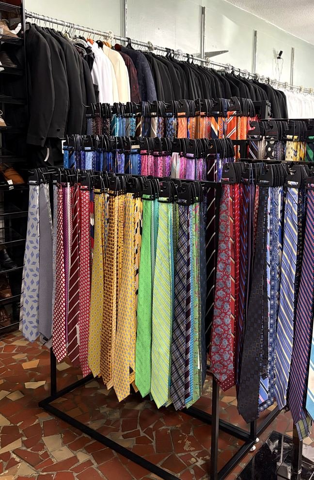 A loaded three-tier rack of neckties in every colour and pattern — stripes, paisley, solids and neats — with the black formalwear rack behind.